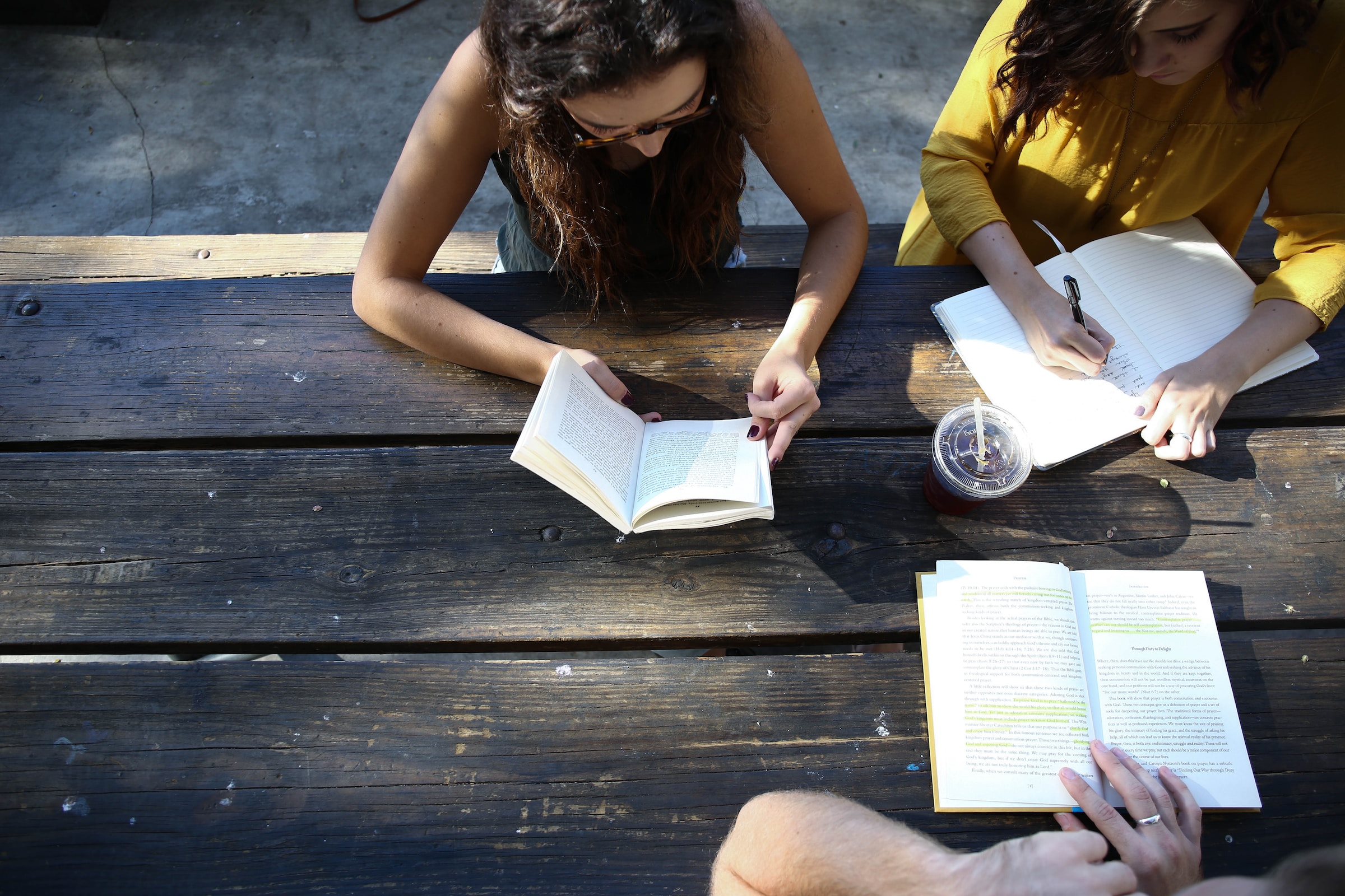 In an aerial view, three students are reading books while sitting at an outdoor picnic table on a sunny day.