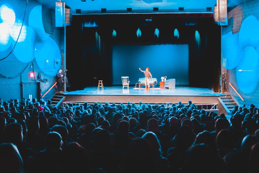 Students perform on stage for a theater production, bathed in blue light signifying night time, in front of a crowded audience