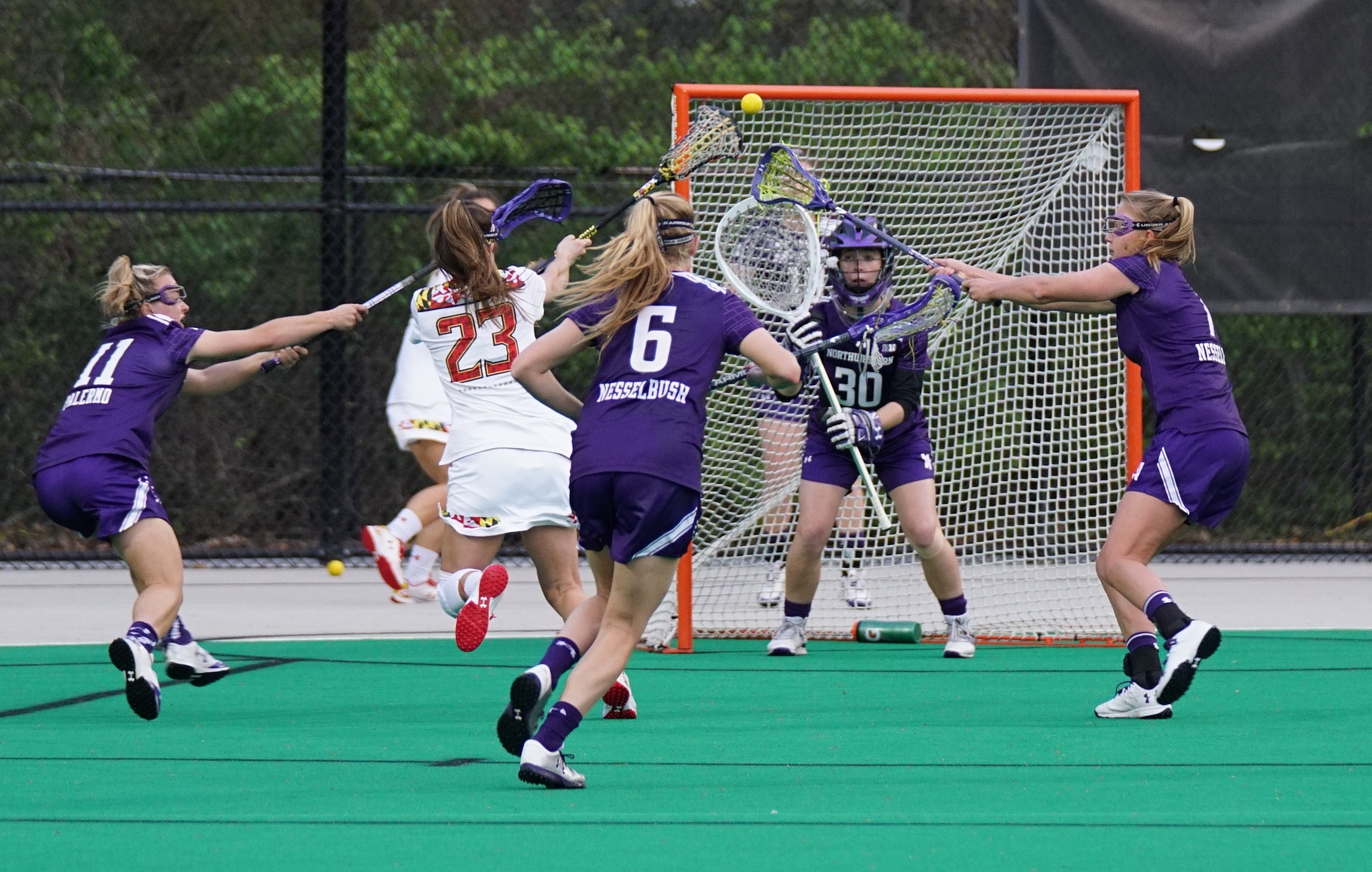 Two women's lacrosse teams compete during playoffs, playing on the field, while the goalie blocks the net.