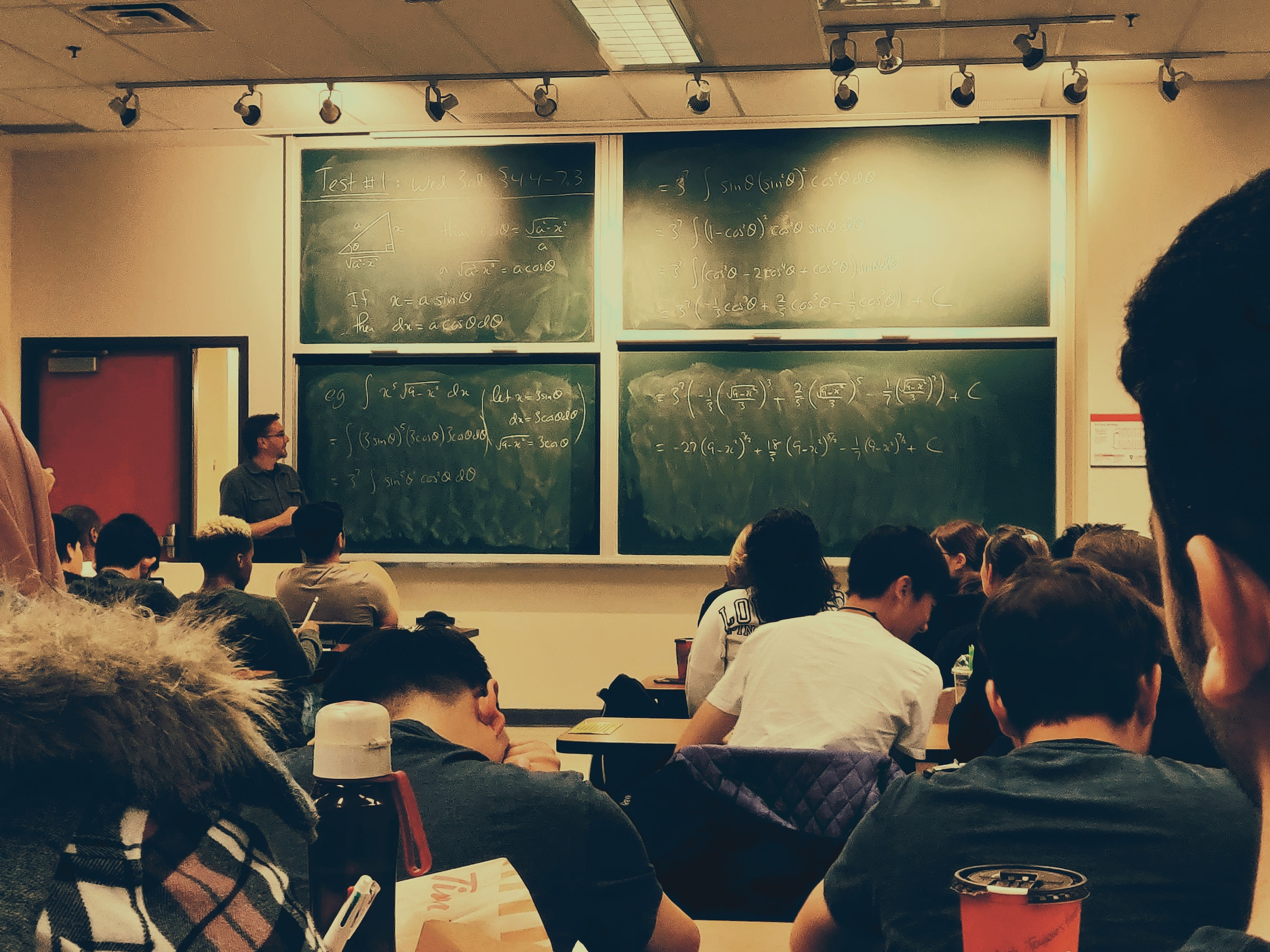 Students sit in rows of chairs in a classroom while the professor explains math concepts that have been written on the board.